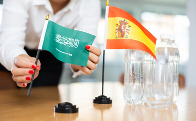 Little flag of Spain on table with bottles of water and flag of Saudi Arabia put next to it by positive young woman 