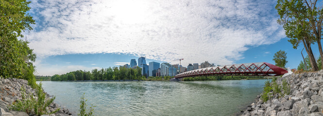 Panoramic skyline view of Calgary, Alberta with Peace Bridge in view over the Bow River in summer spring time on blue sky day with partial clouds. 