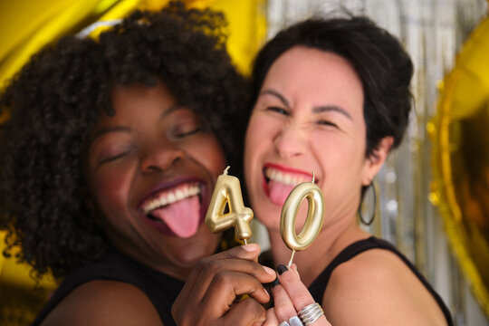 Two multiracial happy women in a 40 birthday party with 40 birthday golden candles sticking out the tongue.