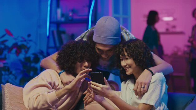 Medium shot of diverse male and female friends sitting together on couch at home party, using smartphone, then young man walking up, hugging them, talking group selfie, looking at photo and smiling