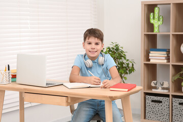 Boy writing in notepad near laptop at desk in room. Home workplace