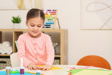 Cute little girl making colorful paper card at desk in room. Home workplace