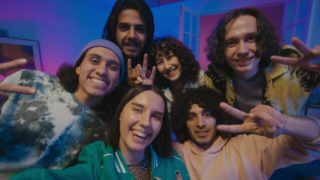 Medium close-up shot of young multiracial couple sitting on couch at house party, beckoning friends, then all taking group selfie and making funny faces and hand gestures