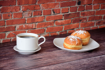 A cup of coffee on a saucer with a spoon and a plate with fresh donuts in powdered sugar on a wooden table against a brick wall.