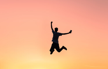 Silhouette of a man holding a smartphone jumping up to celebrate success, with sunset sky in the background. Motivation to achieve success.