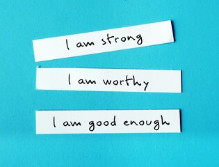 Three note paper on blue background with handwritten text I AM STRONG, I AM WORTHY, I AM ENOUGH -...