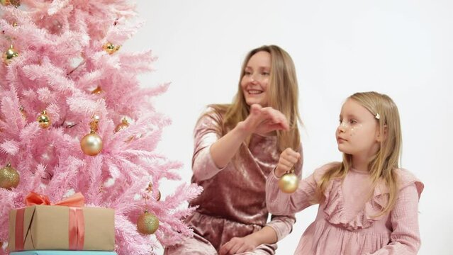 Mother and little daughter sit next to pink Christmas tree and decorating it with golden ball. Happy family hang balls on Ney Year fir and then hugging. Tender relationship between mom and her kid