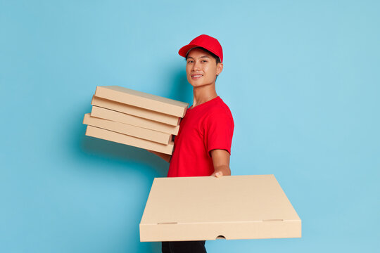 Oriental young guy in red cap and red t-shirt posing against blue wall with pile of pizza boxes, happy guy ready to deliver tasty pizza, food delivery concept, copy space