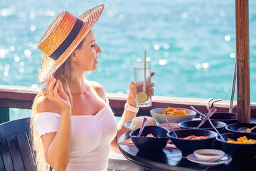 Young woman enjoys a delicious breakfast of exotic fruits and pastries on the balcony of her luxury hotel room. Sun shines brightly and the sea is calm. Girl relaxed and happy, enjoying her vacation.
