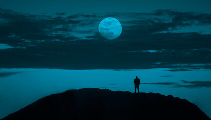 silhouette of one, alone man stands on top of a mountain against fool moon. A male standing on the top of the rock under the clouds. night scene. blue sky.
