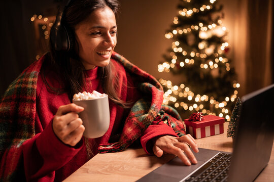Happy woman in headphones using laptop, drinking cocoa, sitting at table near Christmas tree at home, full length. Cheery young lady shopping online, watching Xmas movie