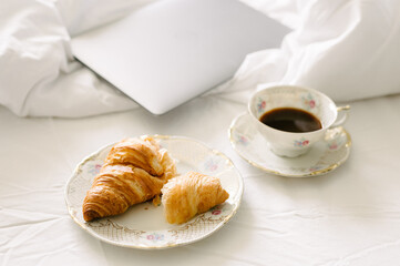 Overhead view of drink served in cup by smart computer laptop, croissant on bed.