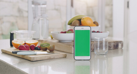 Fototapeta na wymiar Phone, green screen and kitchen with fruit, diet app and table by blurred background in home for smoothie. Smartphone, counter and fruits with space for mock up, logo and nutrition brand in house