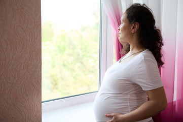 Beautiful pregnant woman future mother holds her belly, looks thoughtfully out the window at home,...