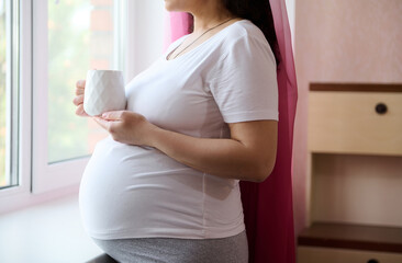 Close-up of a happy pregnant woman in white t-shirt, standing by window at home bedroom, looking...