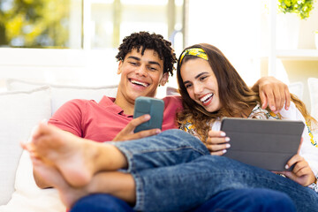 Cheerful biracial young couple using mobile phone and digital tablet while relaxing on sofa
