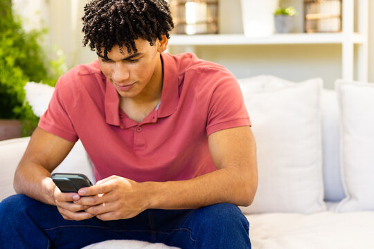 Biracial handsome young man texting over mobile phone while sitting on sofa in living room