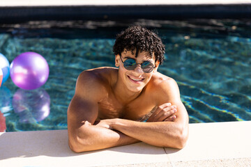 Portrait of biracial shirtless young man in sunglasses smiling and leaning at poolside