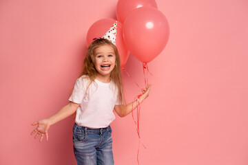 little girl in a white t-shirt and birthday hat holding balloons on pink background