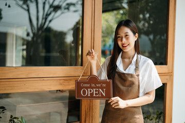 Young female entrepreneur hanging a welcome sign in front of a coffee shop. Beautiful waitress or...