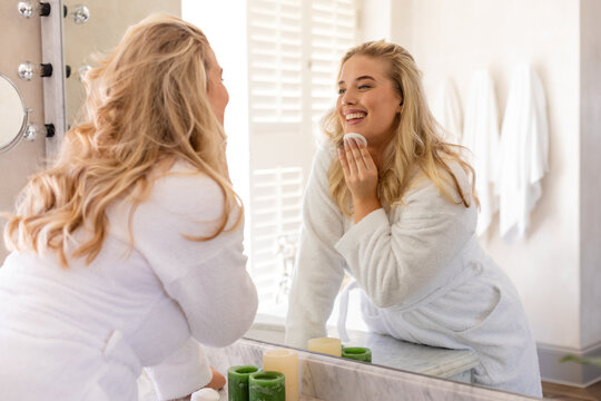 Caucasian plus size woman dressed in bathrobe applying beauty product on face and looking at mirror - Powered by Adobe