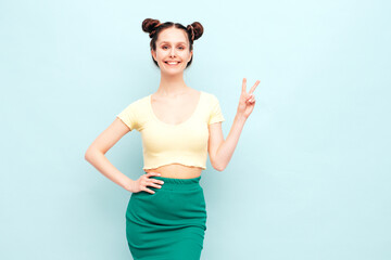 Young beautiful smiling female in trendy summer yellow t-shirt and green skirt. Carefree woman with two horns hairstyle posing near blue wall in studio. Positive model having fun. Cheerful and happy