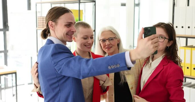 Happy corporate team of company employees standing in modern office and looking at smartphone screen, taking selfie. Friendships at work and modern technology