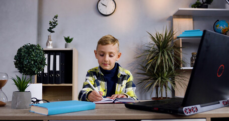 Thoughtful attractive modern boy in plaid shirt sitting at the table and writing notes into the diary