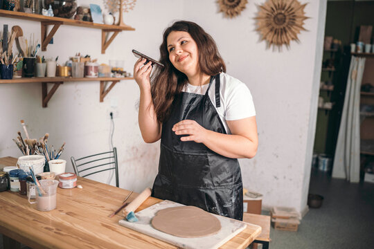 Smiling potter talking on speaker phone standing at workshop