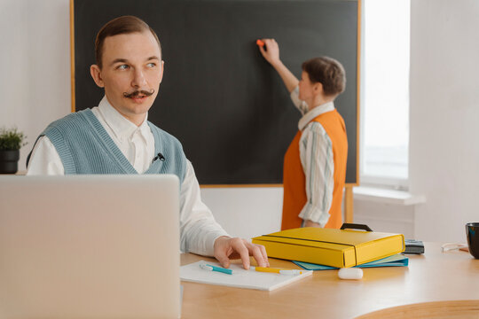 Smiling man sitting in classroom looking at laptop while colleague is writing on blackboard
