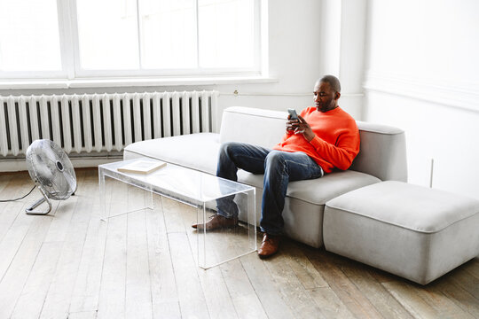 Mature Man Using Smart Phone Sitting In Front Of Fan At Home