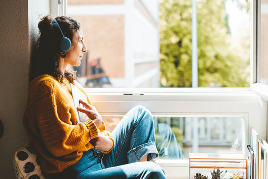 Thoughtful woman wearing headphones sitting near window at home