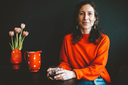 Smiling woman with drink sitting at table with red flower vase in front of black wall
