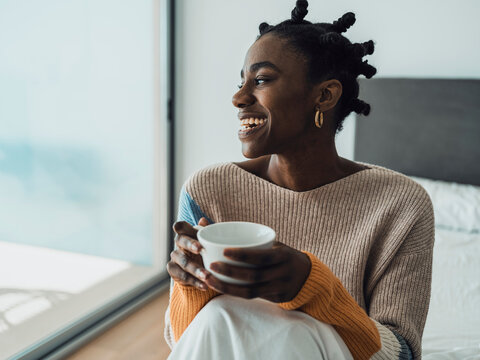 Smiling woman with Afro hairstyle sitting on bed holding coffee cup