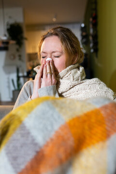 Sick young woman wrapped in blanket sneezing at home