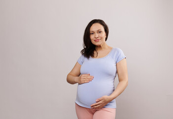 Portrait of beautiful stunning pregnant woman, gravid expectant mother holding palms on her big belly in late pregnancy, smiling looking at camera, standing over isolated background. Maternity concept