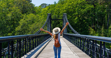 Happy woman tourist with arms raised on suspension bridge over river in the forest- Europa