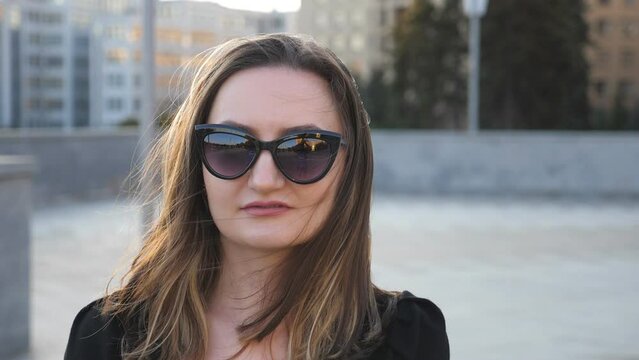 Portrait of young businesswoman in sunglasses with cityscape at background. Attractive business woman looking at camera. Hair of lady fluttering in wind. Face of confident girl. Slow motion close up