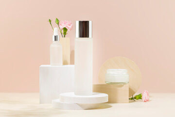 Cosmetic glass jars with natural cream, face serum and organic tonic on podiums on pink background with pink flowers. Organic beauty product and skin friendly cosmetics concept. Mockup, front view