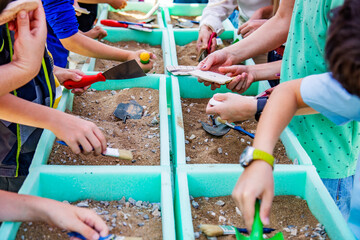 Children working as a archaeologists with brushes and shovels in containers with sand and stones,...
