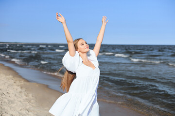 Fototapeta na wymiar Happy smiling beautiful woman on the ocean beach standing in a white summer dress, raising hands.