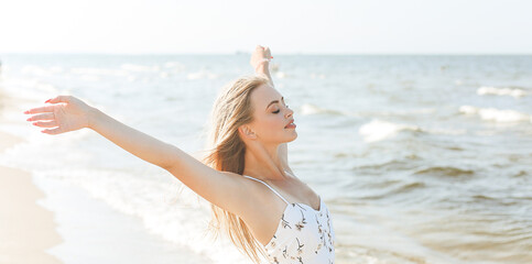 Happy blonde beautiful woman on the ocean beach standing in a white summer dress, open arms.