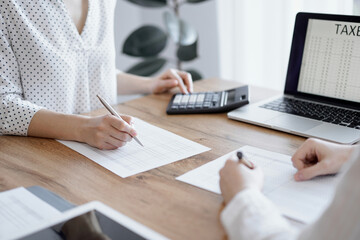 Woman accountant using a calculator and laptop computer while counting taxes with colleague at wooden desk in office. Teamwork in business audit and finance.