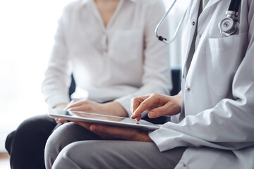 Doctor and patient sitting at sofa in clinic office. The focus is on female physician's hands using tablet computer, close up. Perfect medical service and medicine concept.