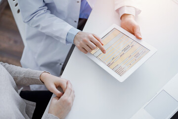 Doctor and patient sitting at the table in clinic. The focus is on female physician's hands using tablet computer, close up. Medicine and healthcare concept.