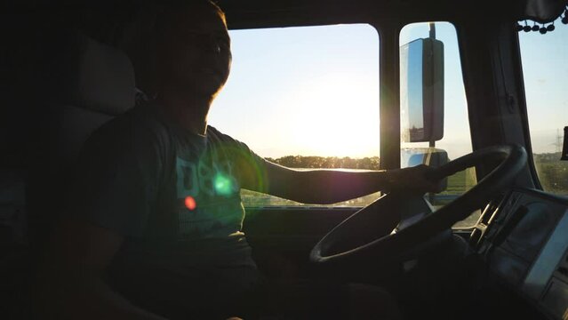 Man is driving a truck and smiling attentively watching the road. Caucasian guy is riding through the countryside on the sunset background. Lorry driver profile.Side view Slow motion Close up