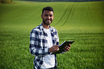 Obraz premium Standing and holding notepad. Handsome Indian man is on the agricultural field