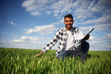 Naklejka na ściany i meble Sitting, checking the wheat, holding notepad. Handsome Indian man is on the agricultural field
