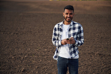 Standing and holding smartphone. Cultivated field. Handsome Indian man