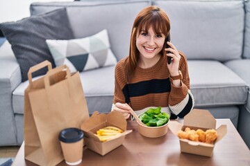 Young woman talking on the smartphone eating take away food at home